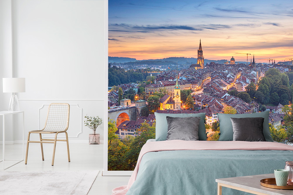 Fototapete - Eine Schlafzimmerwand mit einer großformatigen Fototapete zeigt die Stadt Bern bei Sonnenuntergang, mit Kirchtürmen und warmem Abendlicht.