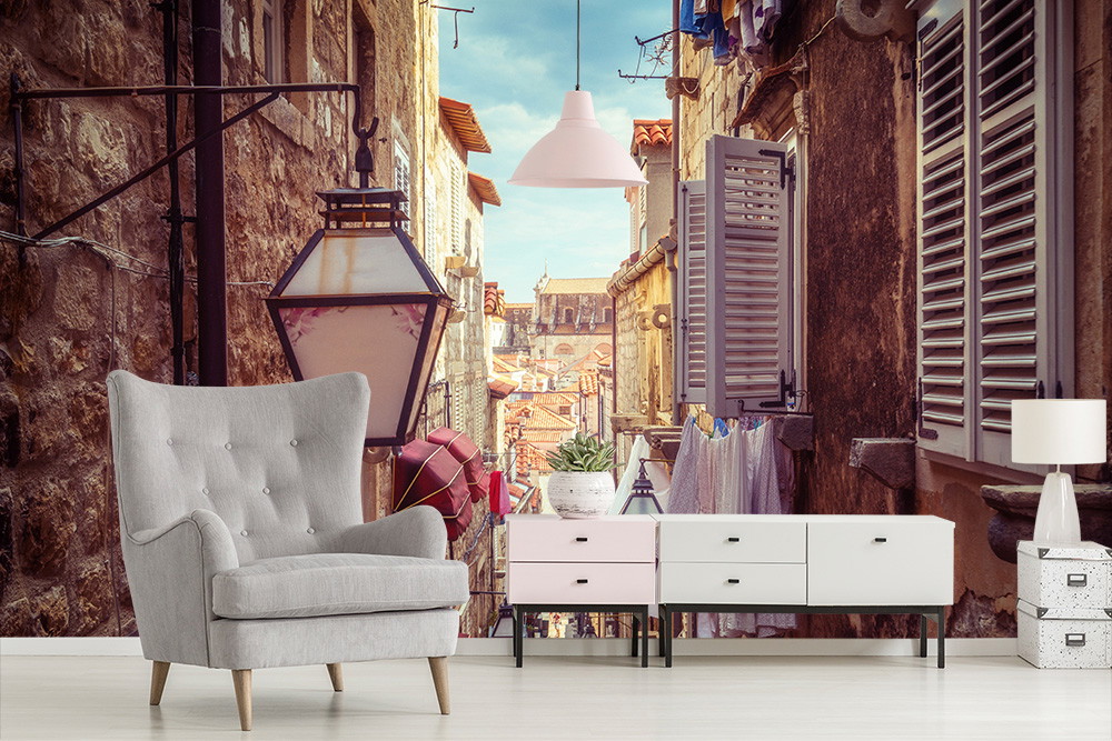 Fototapete - Innenansicht eines gemütlichen Zimmers mit Sessel und Kommode vor einer Wand mit einem Fototapeten-Motiv einer sonnigen Gasse in einer Altstadt