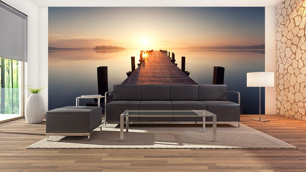Papier peint photo - Panorama d'un lever de soleil sur un lac tranquille avec un ponton en bois, dans un salon moderne avec un canapé gris, un tapis et un lampadaire.