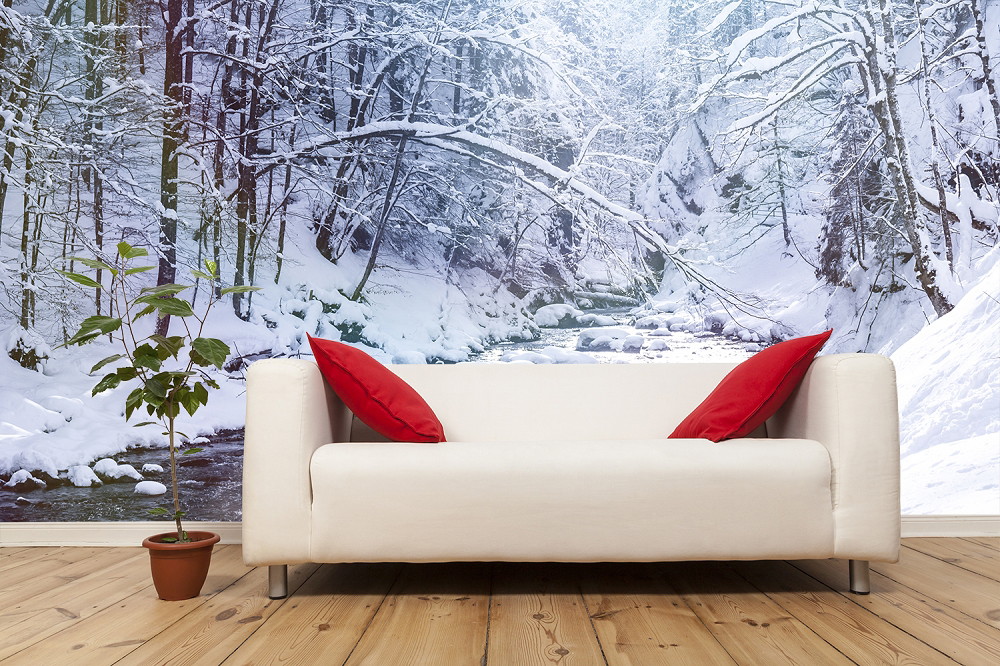 Fototapete - Verschneite Waldszene hinter einem Sofa mit roten Kissen, neben einem Topfpflanze auf Holzboden
