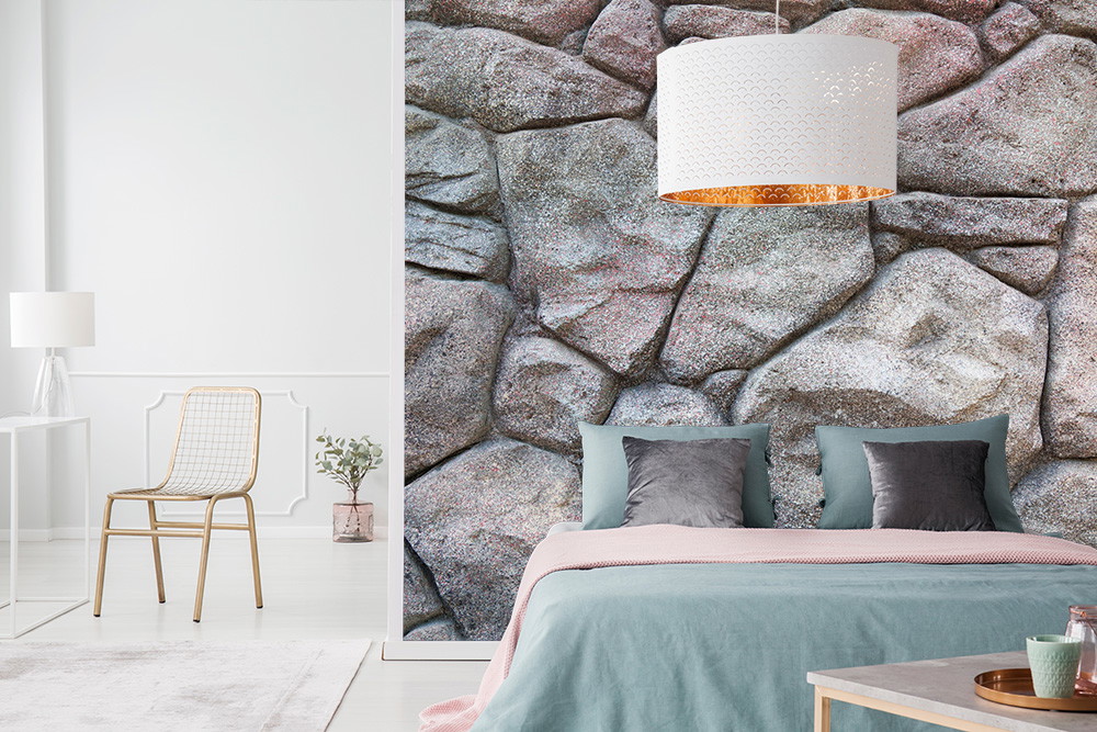 Fotobehang - Slaapkamer met muur in steenlook, grijze kussens, blauw en roze beddengoed en een witte lampenkap