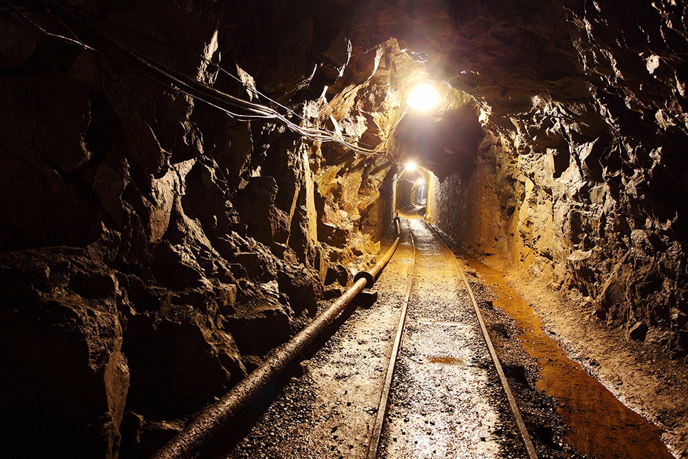 Fototapeta - Oświetlony tunel kopalniany z szynami biegnie przez tunel skalny, ściany z szorstkiej skały otaczają ścieżkę.