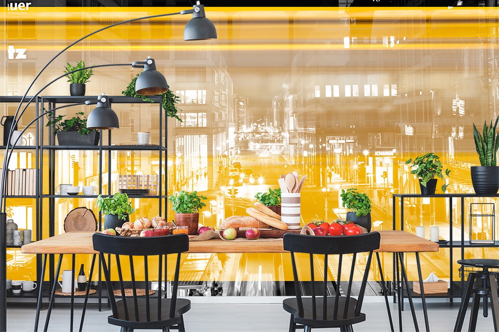 Papier peint photo - Coin repas dans une cuisine avec un motif de ville sur le mur, une table remplie de nourriture et des plantes tout autour